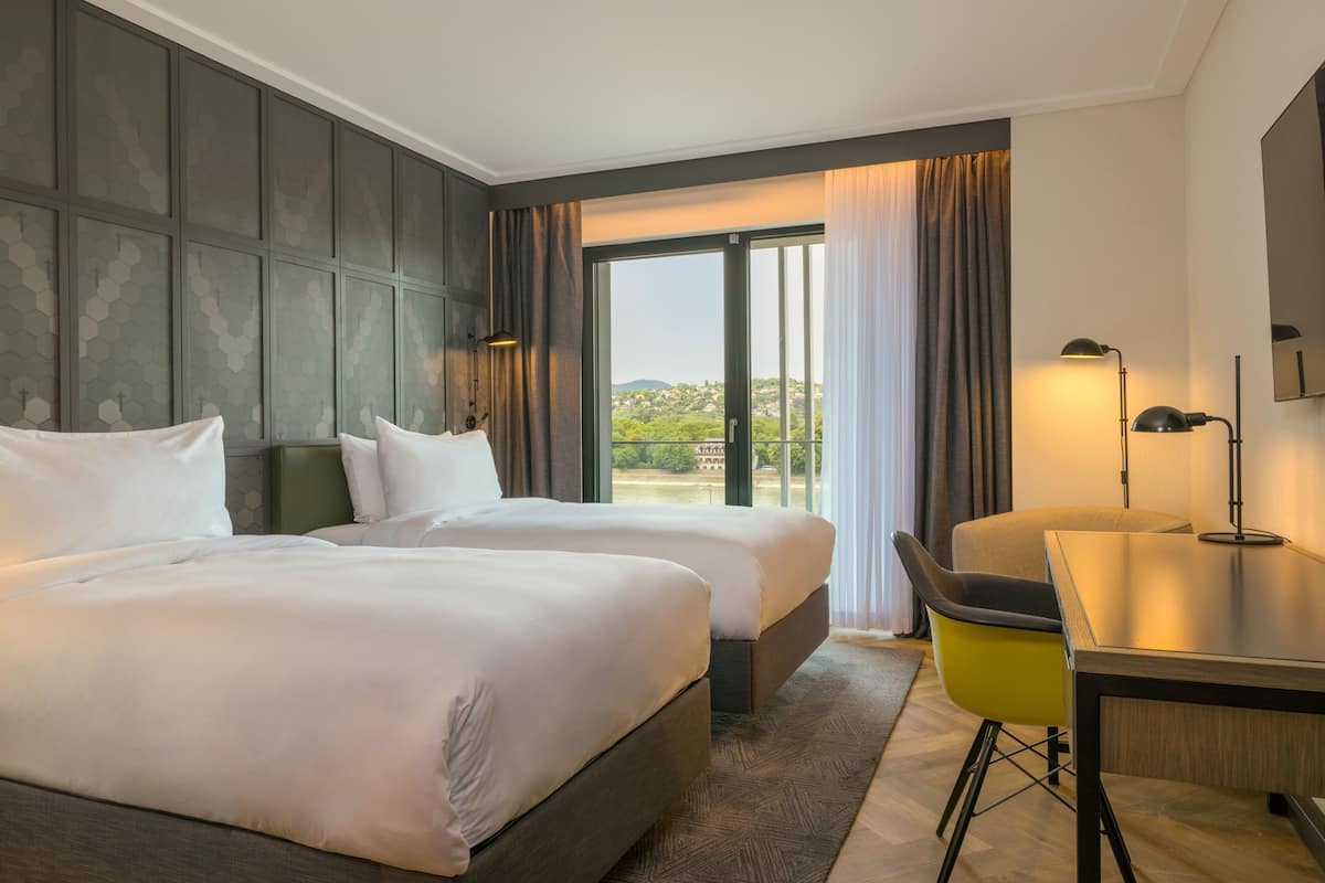 A hotel room with two beds, a desk, a chair, and a large window with a view of the landscape.