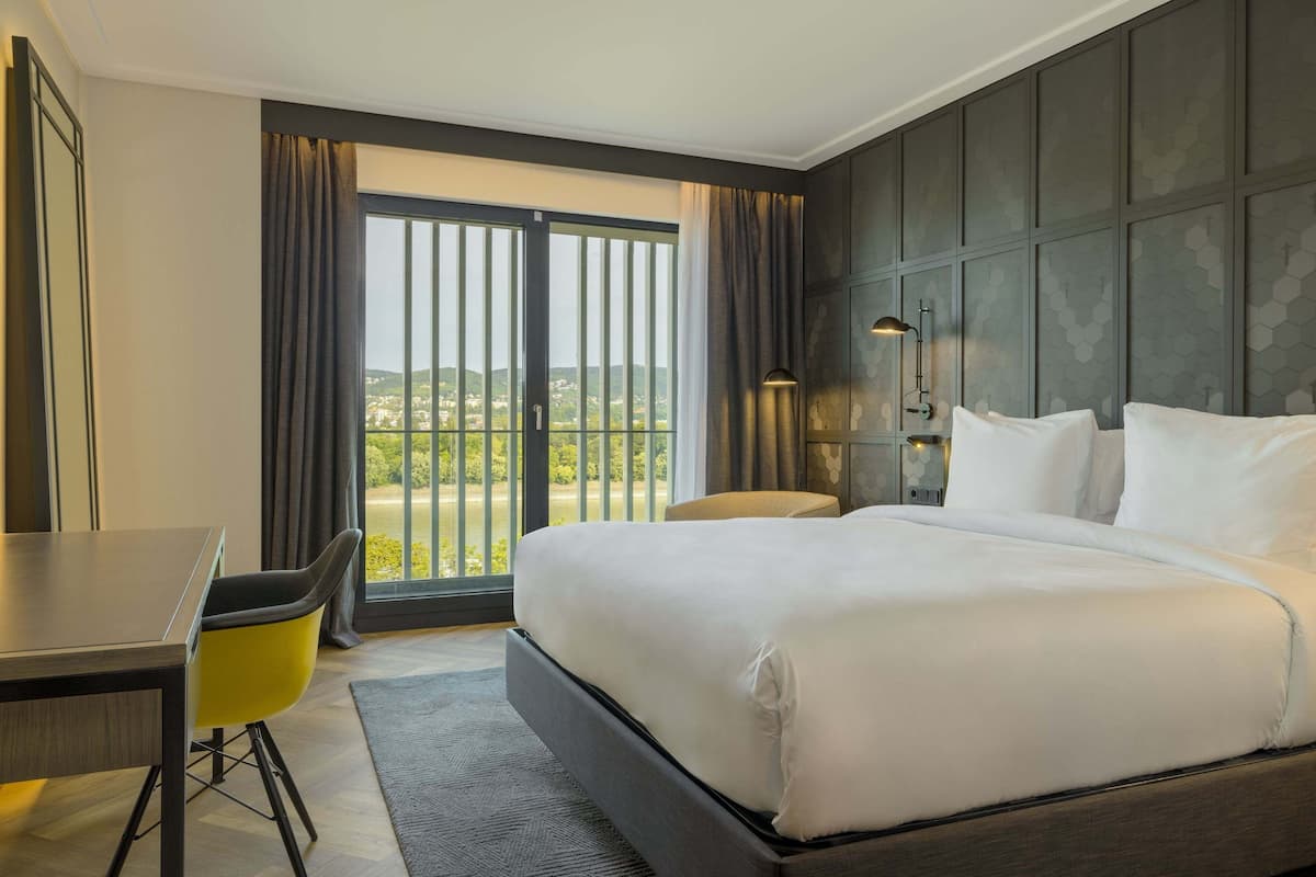 A hotel room with a bed, a desk, a chair, and a view of the outdoors through a large window.