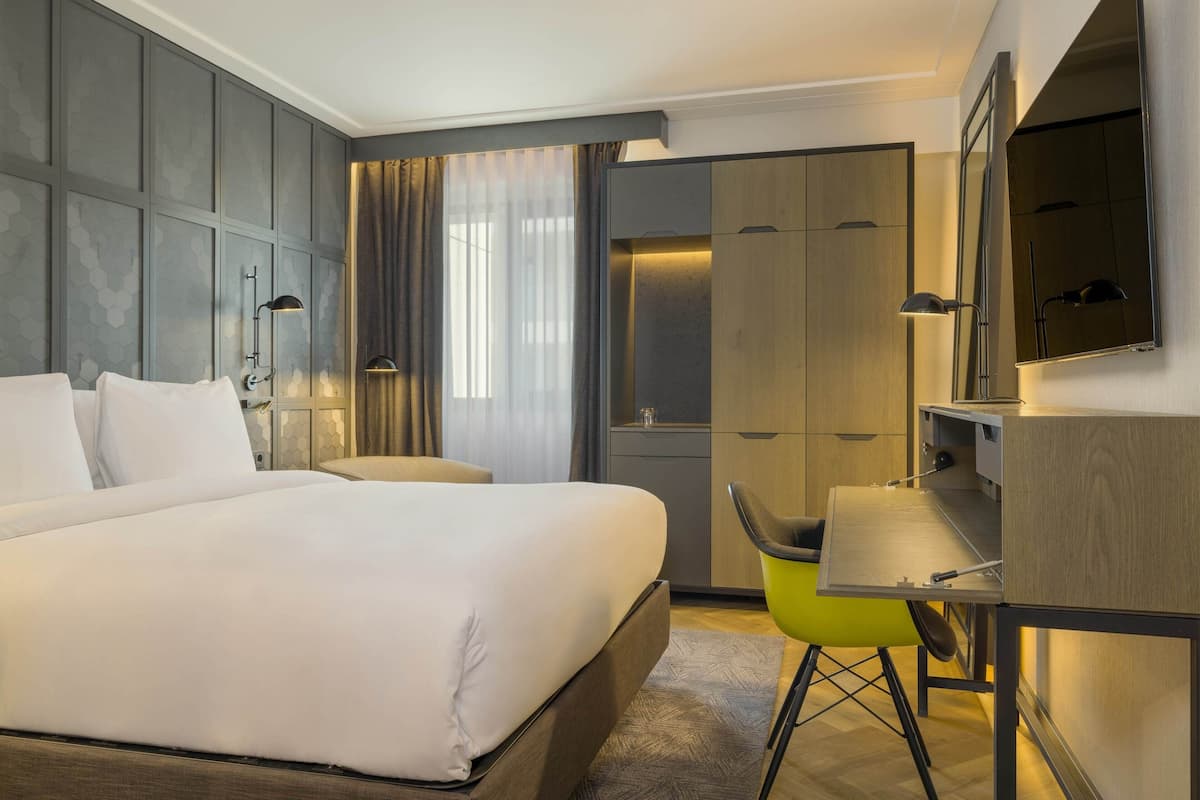 A modern hotel room with a large bed, a desk, a chair, and a wardrobe.