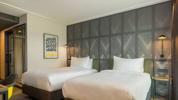 A hotel room with two beds, a desk, a chair, and a wall-mounted lamp.