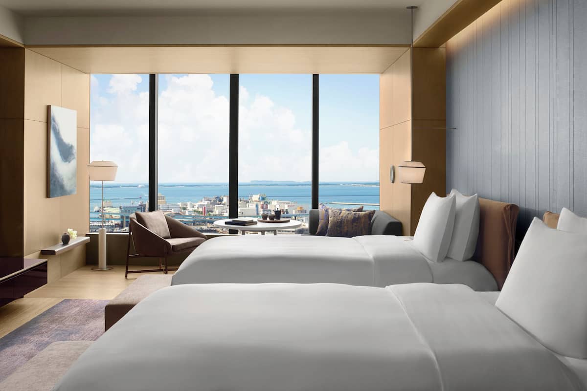 A hotel room with two beds, a sitting area, a view of the city, and a large window.