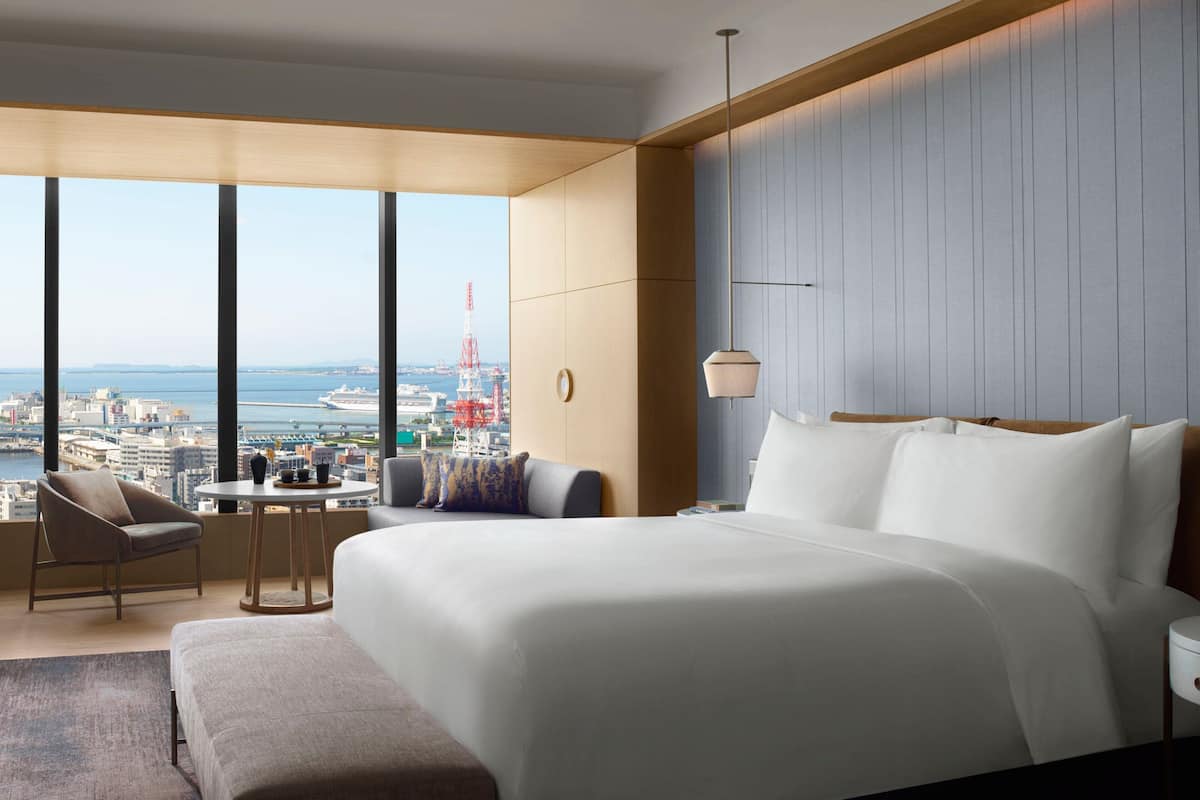 A modern hotel room with a large bed, a seating area with a table, and a view of the cityscape and water.
