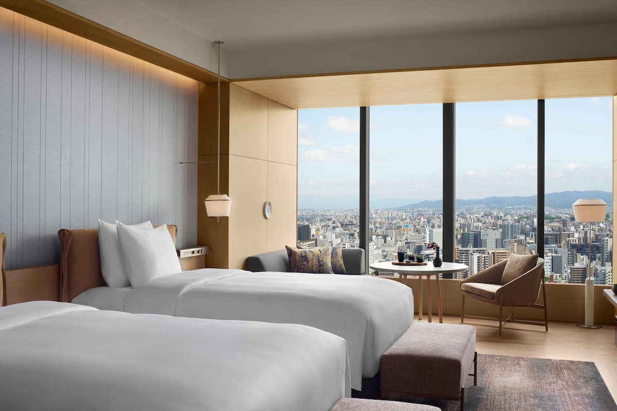 A modern hotel room with two beds, a small table, a chair, and a large window offering a city view.