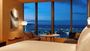 A hotel room with a large window view of a cityscape at dusk, a bed, a chair, a small table with a lamp, and a painting on the wall.