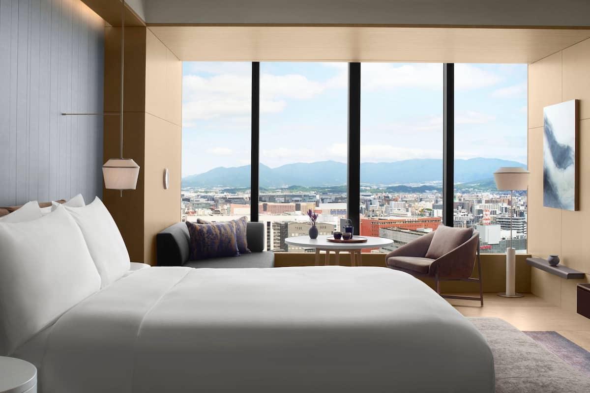 A modern hotel room with a large bed, a seating area with a table, and a city view through large windows.