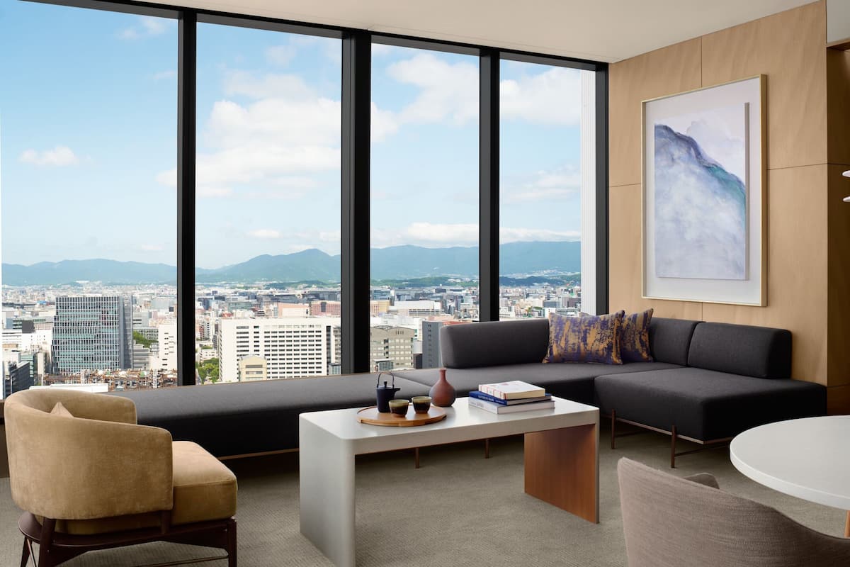 A modern living room with a sofa, armchair, coffee table, and a city view through large windows.