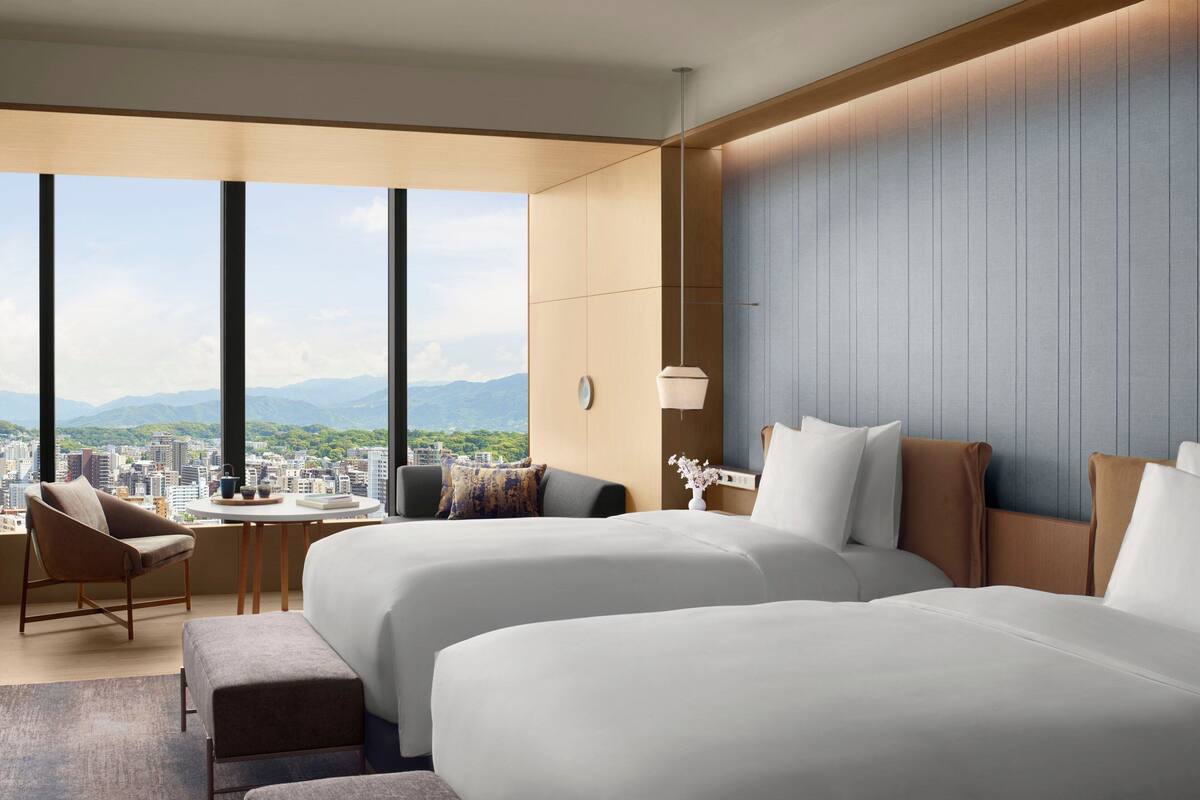 A hotel room with two beds, a sitting area with a chair and table, and a view of a cityscape and mountains.