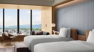 Une chambre d’hôtel avec deux lits, un coin salon comprenant une chaise et une table, et une vue sur le paysage urbain et les montagnes.