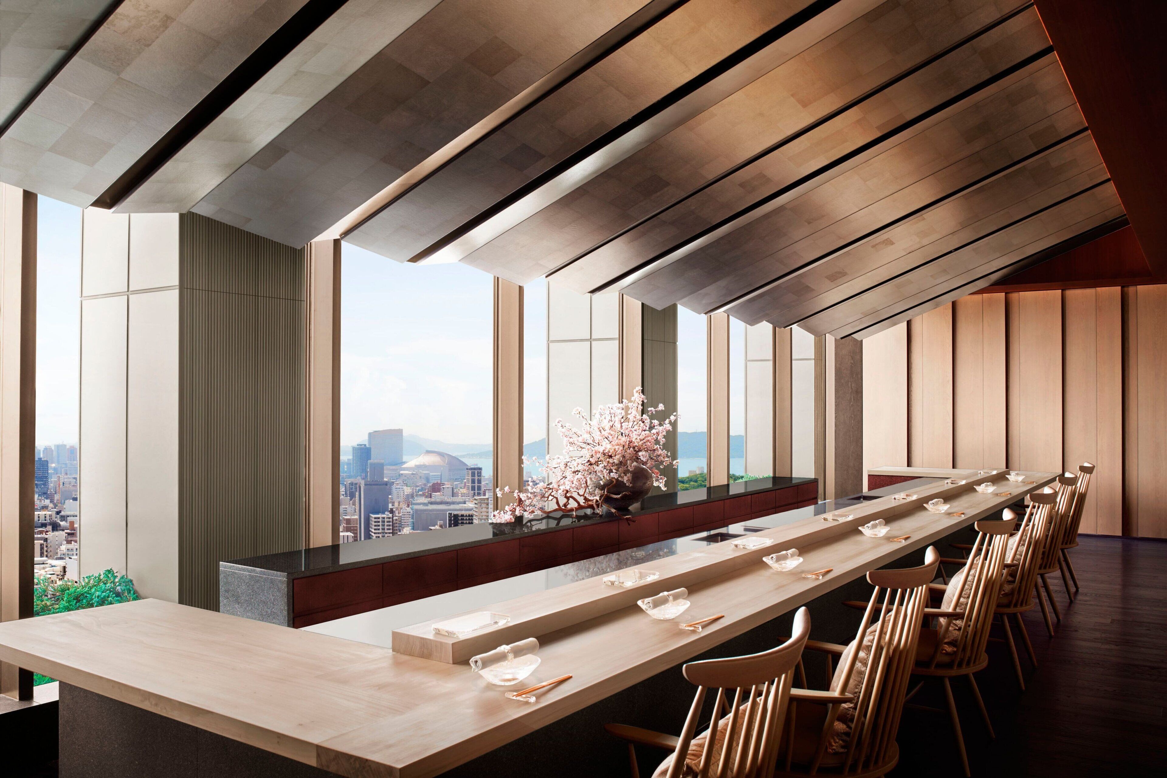 A modern dining area with a long table, chairs, and a city view through large windows.