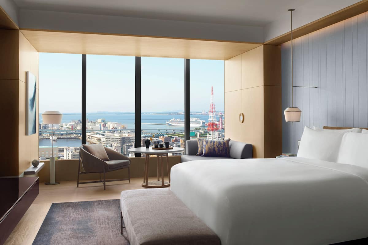 A modern hotel room with a large bed, a seating area with a table, and a view of the cityscape and water.