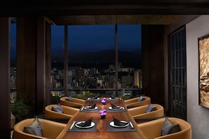 A dining area with a long table set for six, surrounded by cushioned seating, with a cityscape view through large windows.