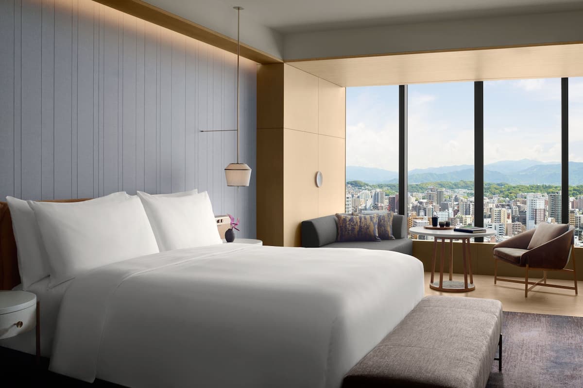 A modern hotel room with a large bed, a seating area with a table, and a view of a cityscape through large windows.
