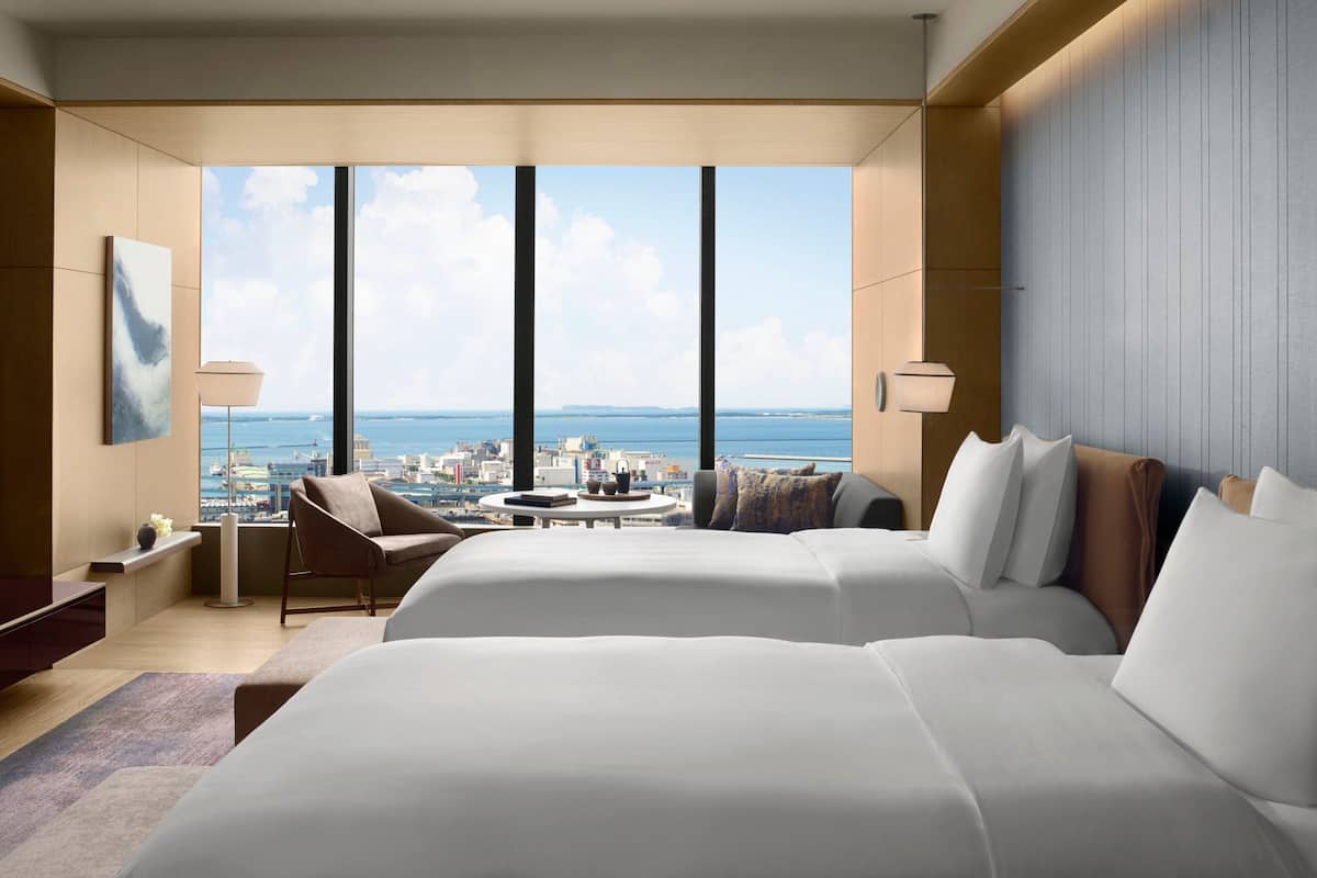 A modern hotel room with two large beds, a sitting area with a chair and table, and a large window offering a city and sea view.