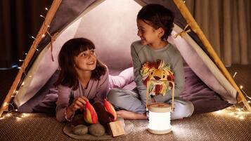 Two children sitting inside a tent with fairy lights, surrounded by toys and a lantern.