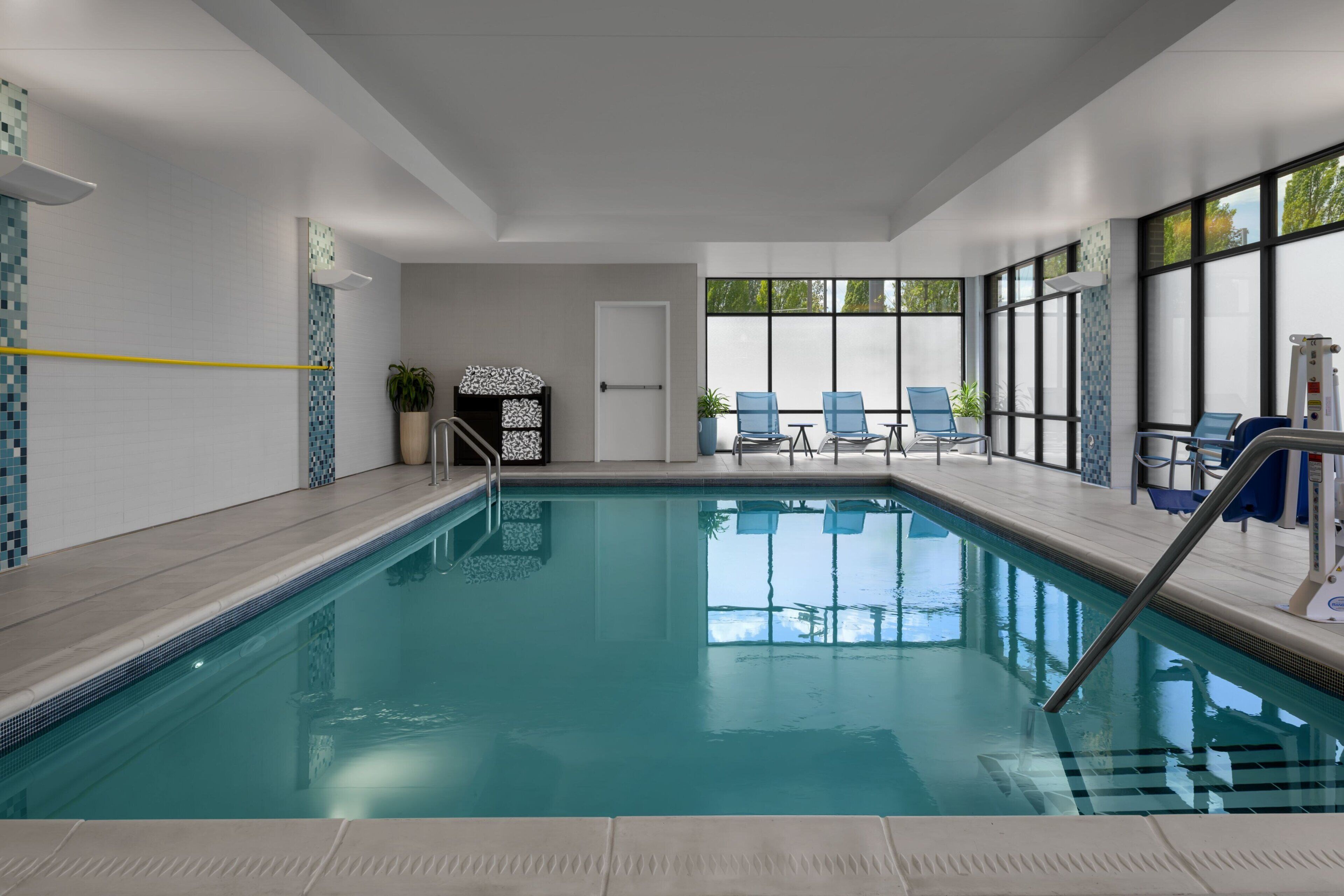 An indoor swimming pool with clear water, a tiled deck, and blue chairs by the windows.