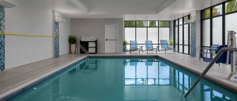 An indoor swimming pool with clear water, a tiled deck, and blue chairs by the windows.