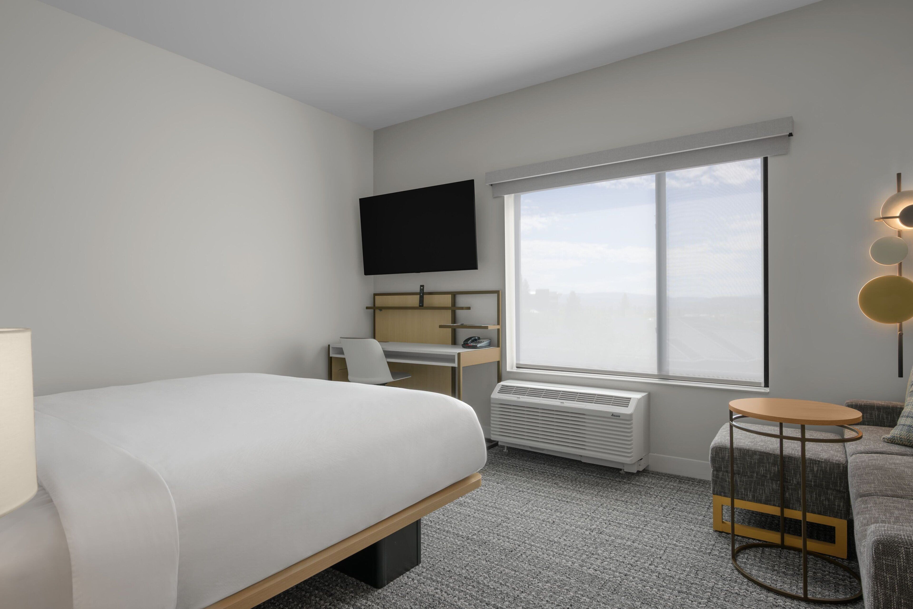 Una habitación de hotel moderna con cama, escritorio, silla y un ventanal grande.
