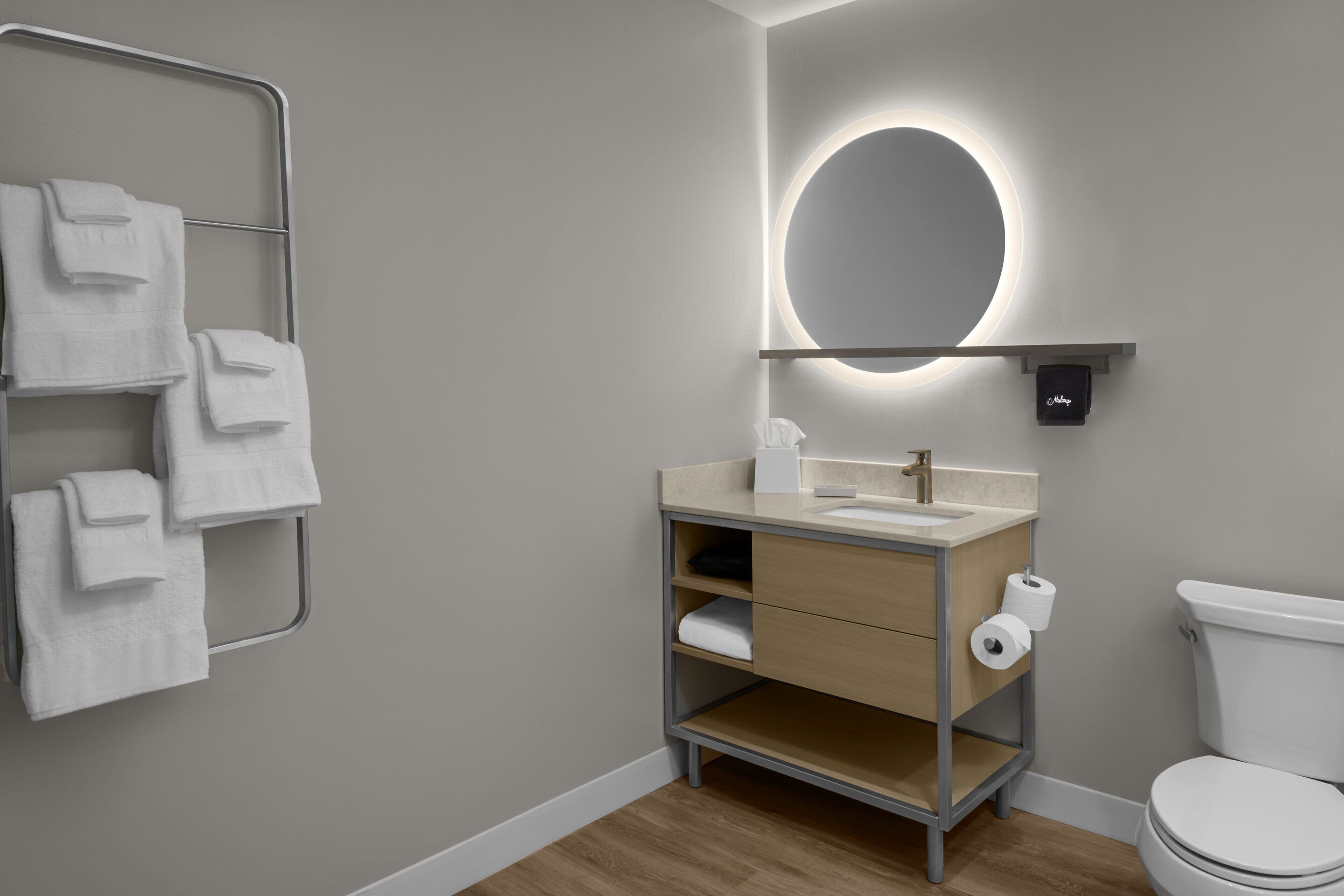 Une salle de bain moderne avec un meuble-lavabo en bois, un miroir rond avec éclairage intégré, un porte-serviettes et des toilettes.