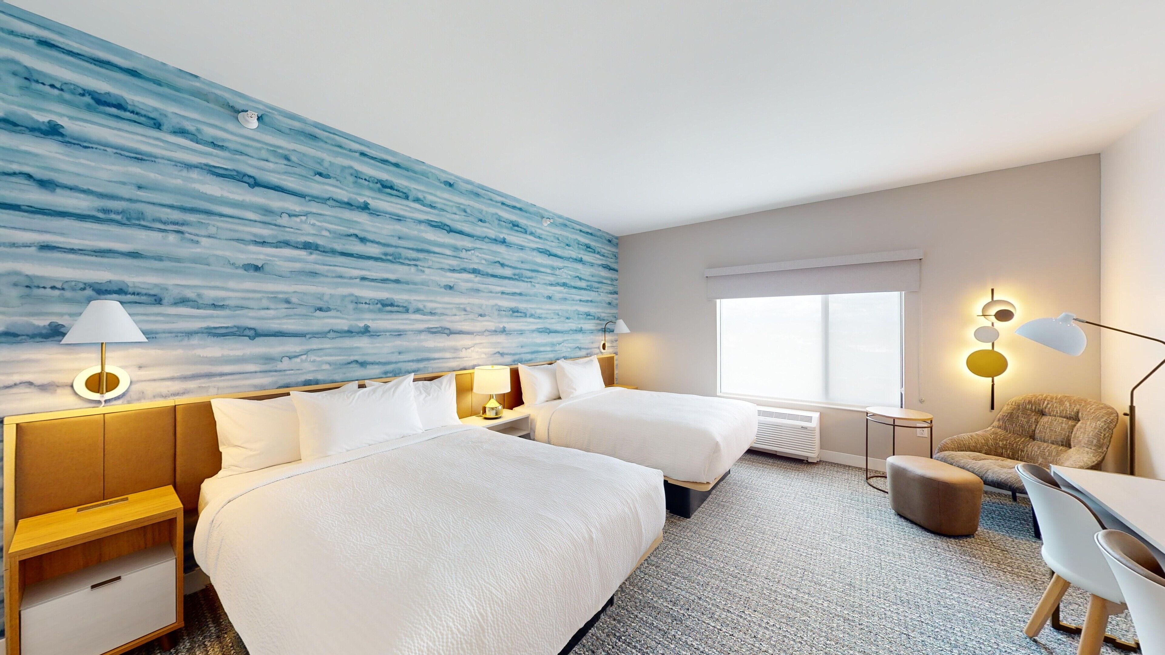 Quarto de hotel com duas camas, uma escrivaninha, uma cadeira, um abajur e uma janela grande.