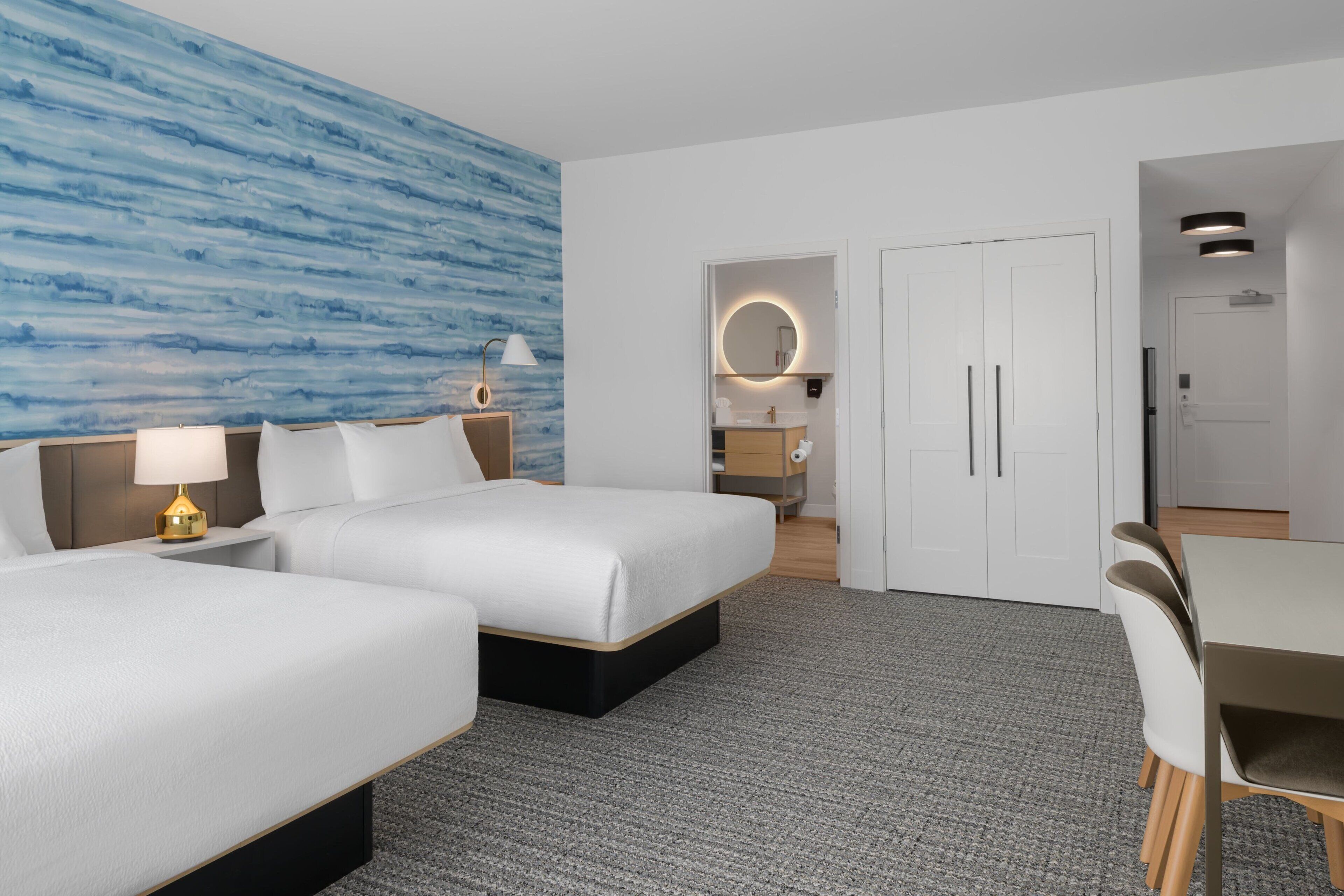 A hotel room with two beds, a desk, a chair, a lamp, a mirror, and a bathroom visible through an open door.