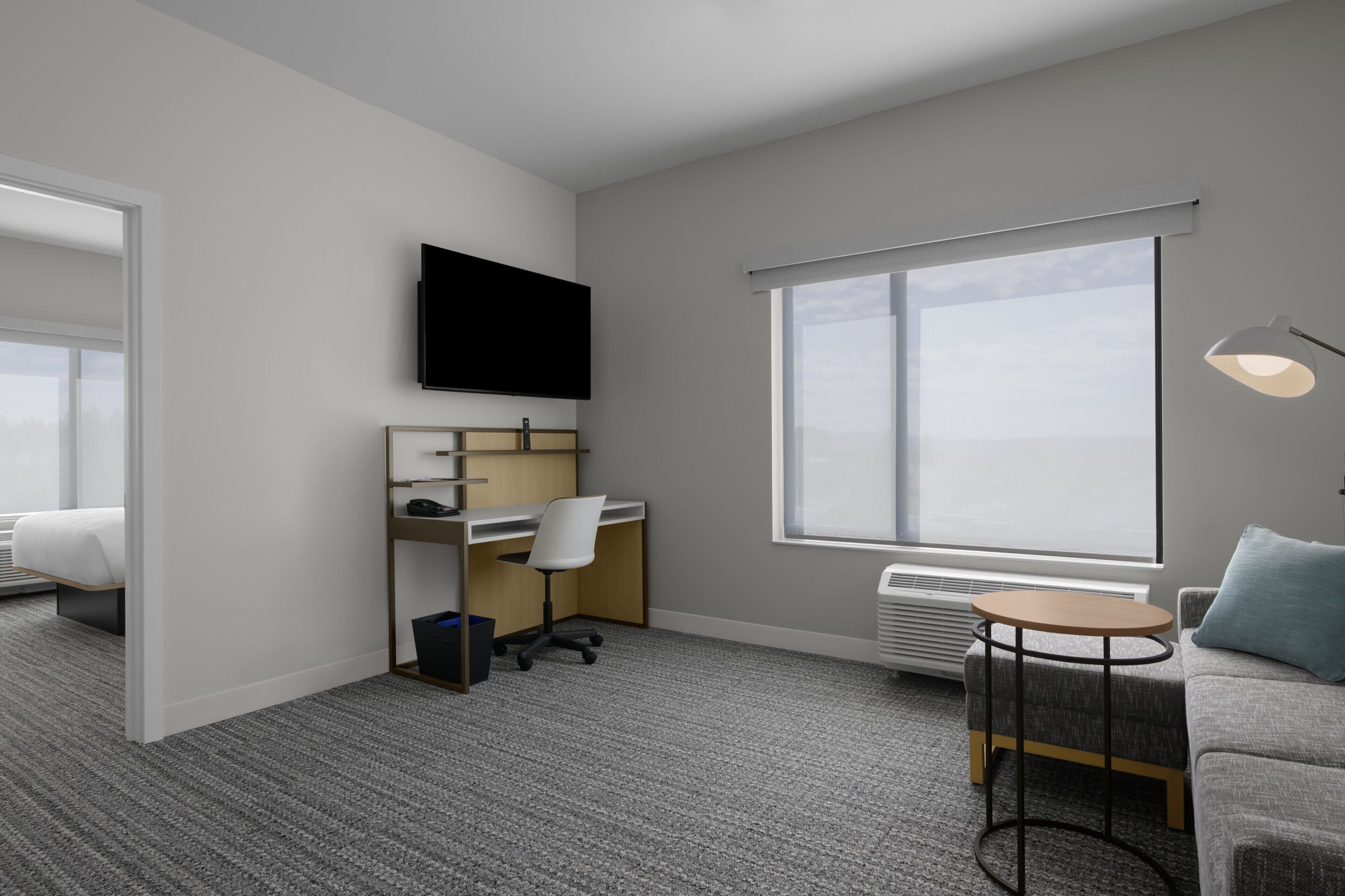 A modern hotel room with a bed, a desk, a chair, a TV, a large window, and a small table with a lamp.