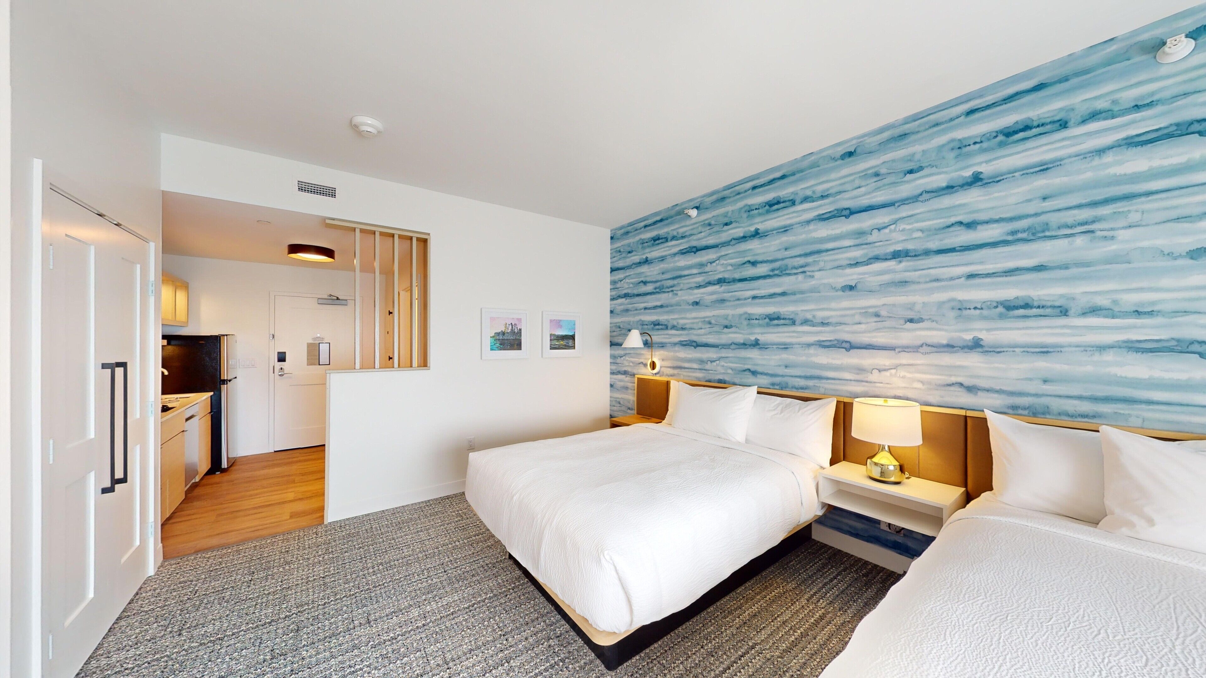 A hotel room with a bed, bedside tables, a nightstand, a wall with a blue abstract design, a door leading to a kitchen, and a hallway.