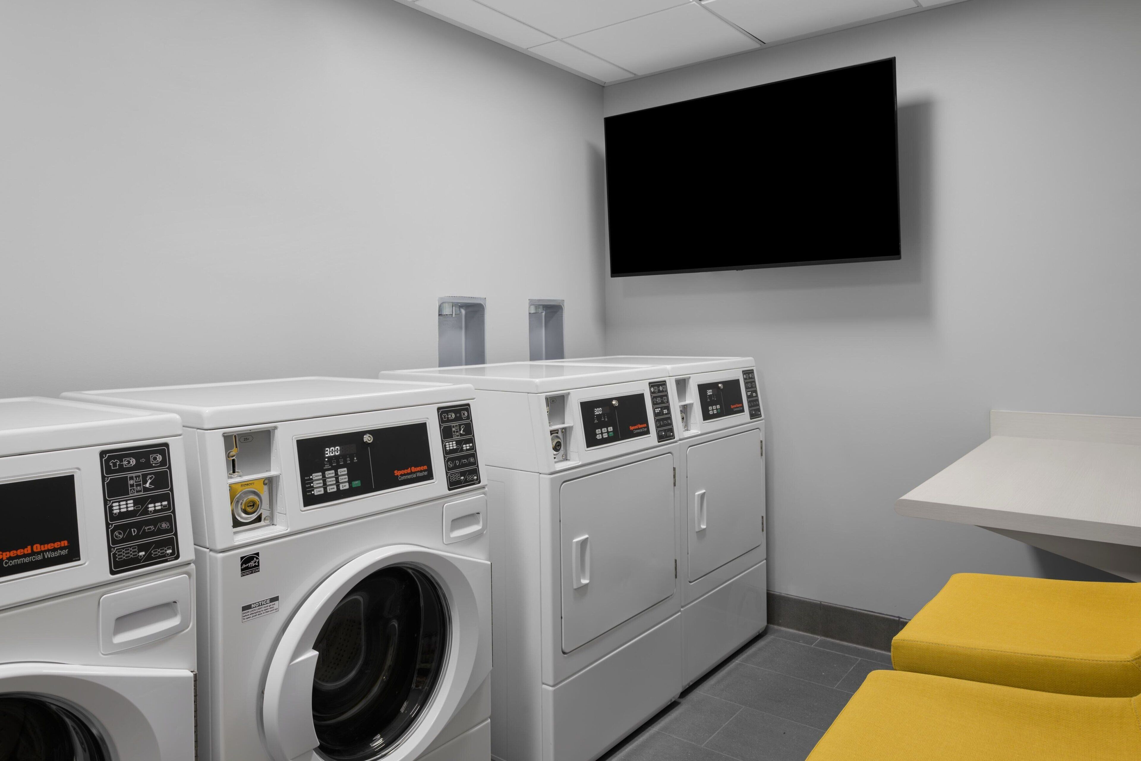A laundry room with multiple washing machines and dryers, a flat-screen TV mounted on the wall, and a small yellow bench.