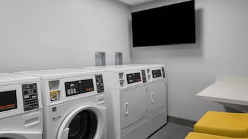 A laundry room with multiple washing machines and dryers, a flat-screen TV mounted on the wall, and a small yellow bench.