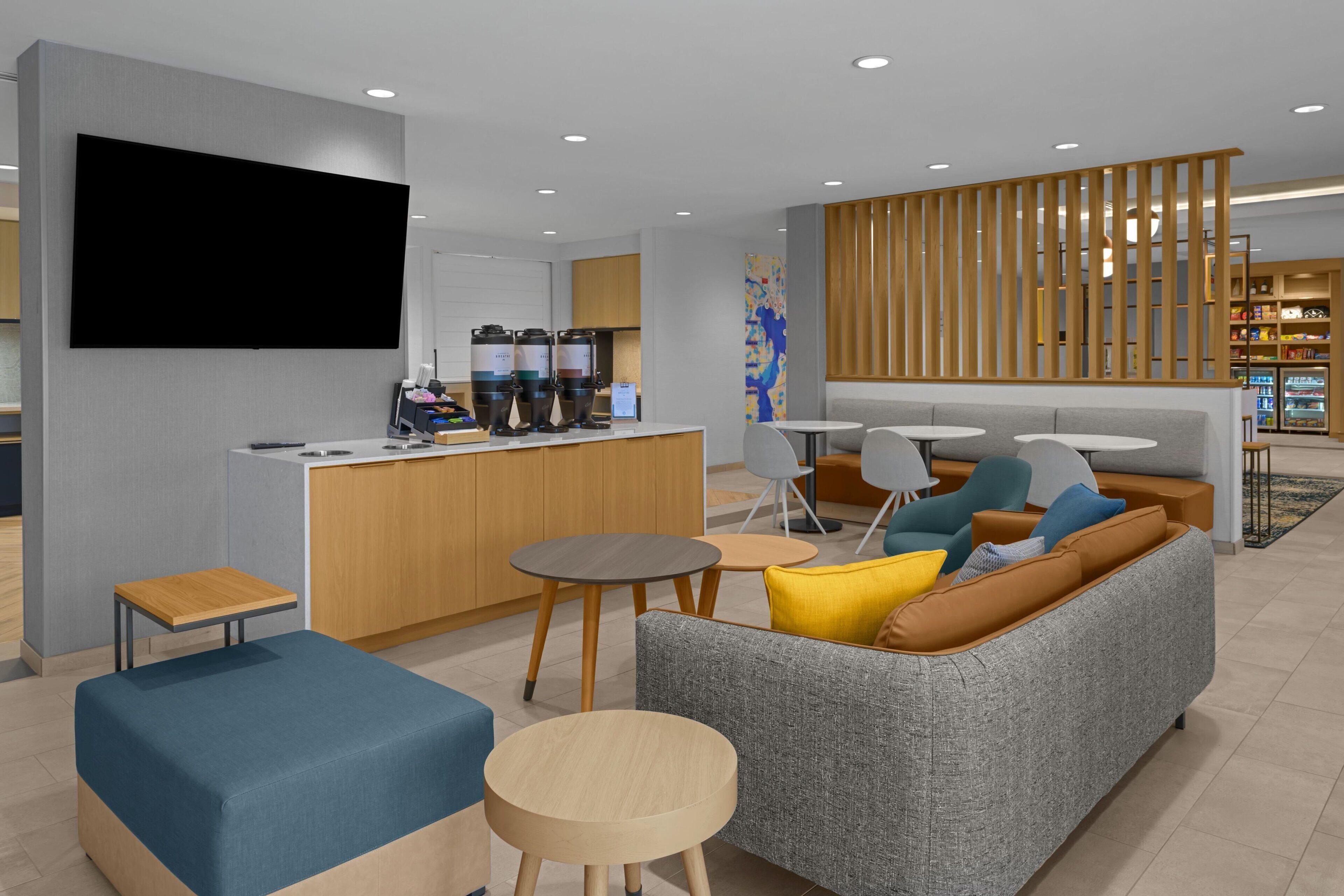A modern hotel lobby with a flat-screen TV, a bar area with coffee machines, a grey sofa with colorful cushions, and a wooden partition.