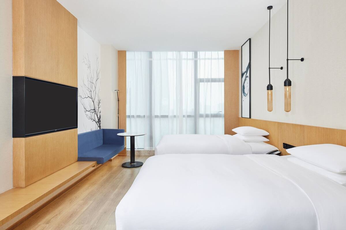 A modern hotel room with a large bed, a TV mounted on a wooden panel, a blue sofa, a small round table, and a large window with sheer curtains.