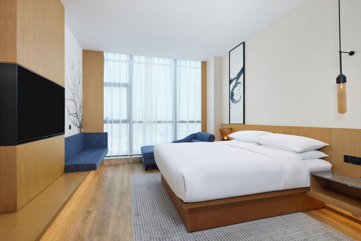 A modern hotel room with a large bed, a flat-screen TV, a sofa, and a large window with sheer curtains.