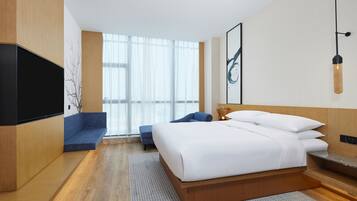 A modern hotel room with a large bed, a flat-screen TV, a sofa, and a large window with sheer curtains.