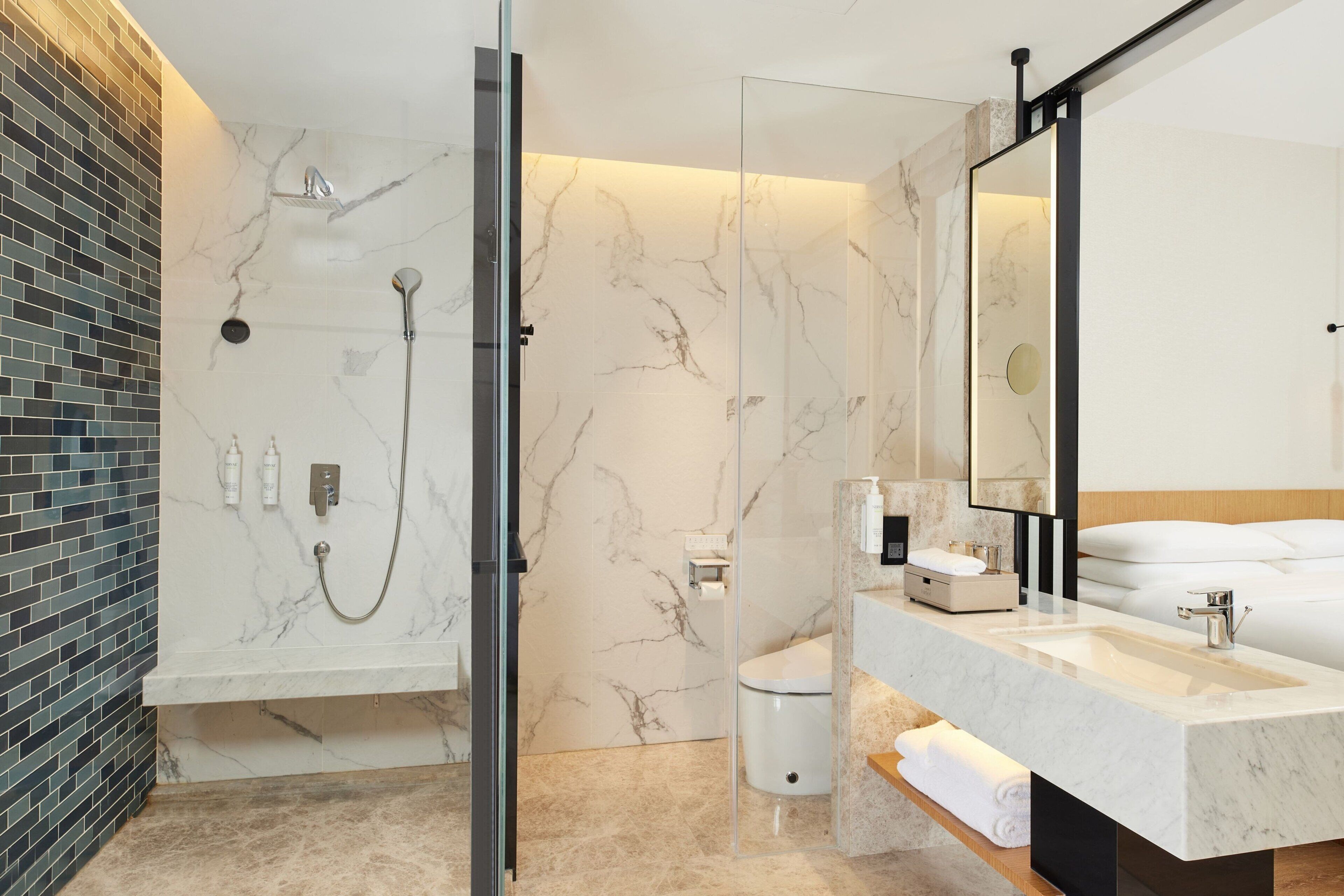 Un baño moderno con ducha de vidrio, pared de azulejos de mármol, espejo grande y lavamanos blanco.