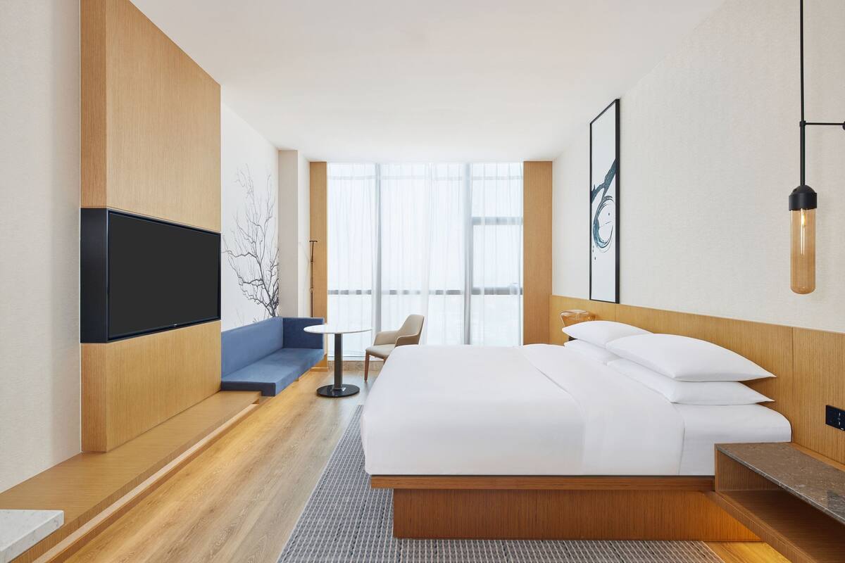 A modern hotel room with a large bed, a flat-screen TV mounted on the wall, a seating area with a sofa and armchair, and a large window providing natural light.