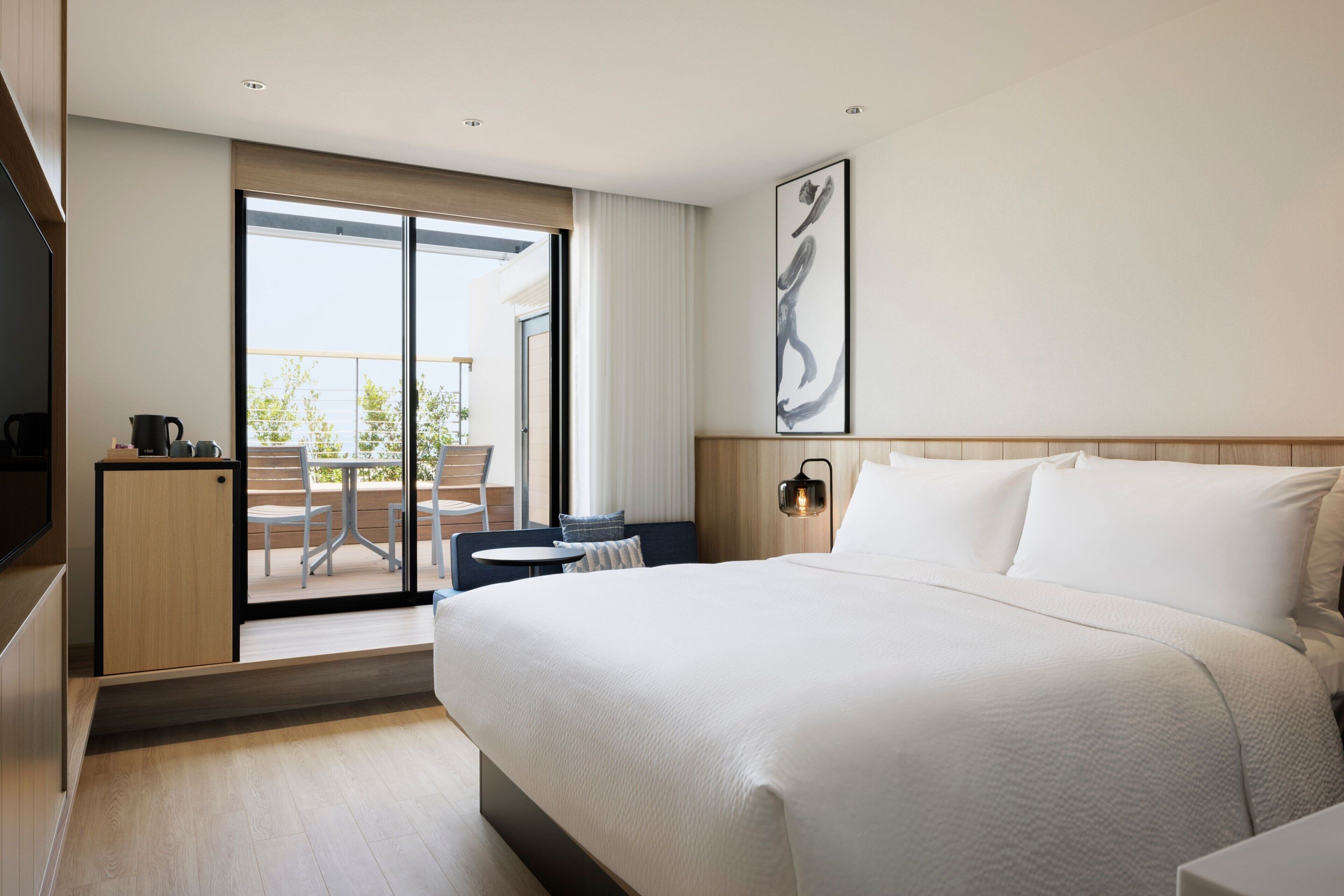 Een moderne hotelkamer met een groot bed, een bureau met stoel, een klein kitchenette en een glazen schuifdeur naar een buitenruimte.
