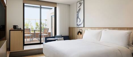 A modern hotel room with a large bed, a desk with a chair, a small kitchenette, and a sliding glass door leading to an outdoor area.