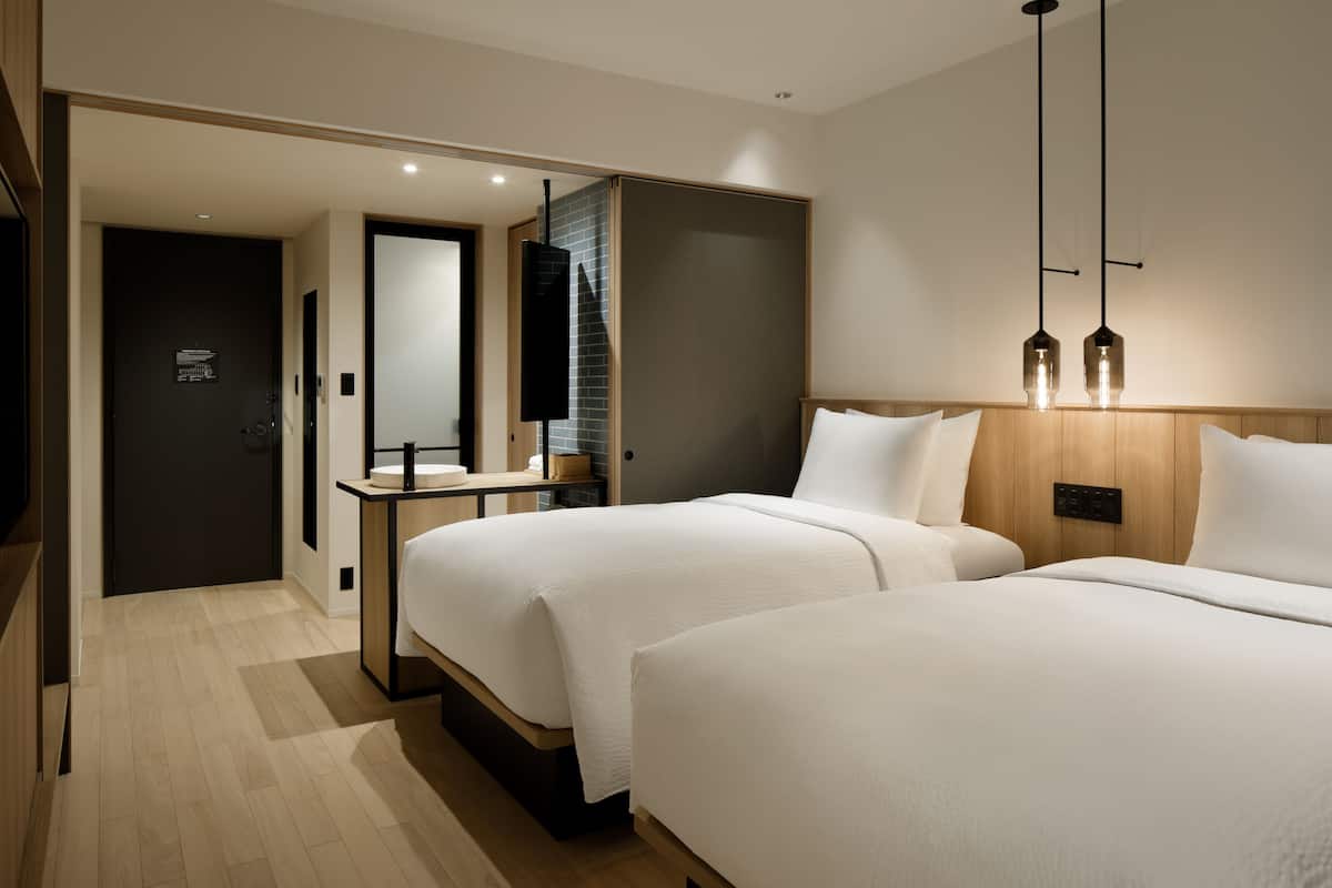 A modern hotel room with two beds, a wooden headboard, a bedside table, a mirror, and a hanging light fixture.