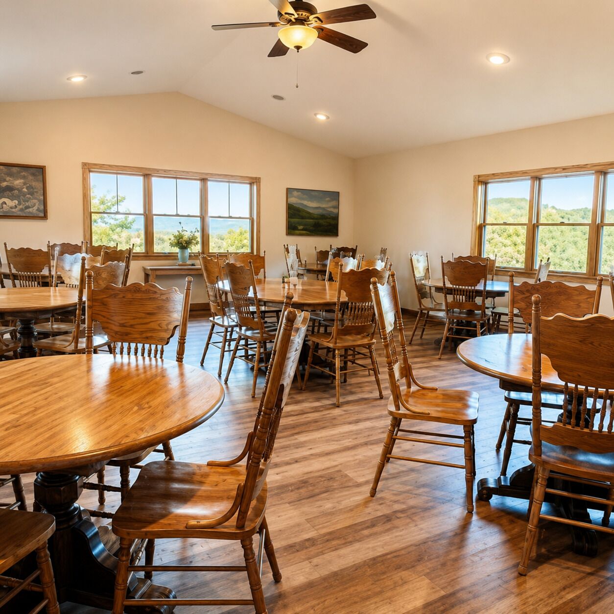 A dining area with multiple wooden tables and chairs, large windows with a view, and a ceiling fan.