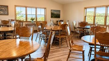 A dining area with multiple wooden tables and chairs, large windows with a view, and a ceiling fan.