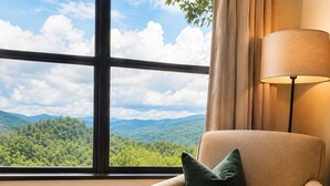 Quarto com uma grande janela que oferece vista para uma cadeia montanhosa arborizada.