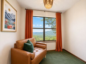Una habitación con un sofá de cuero marrón, un cojín verde, un cuadro enmarco de un paisaje, cortinas naranjas y vistas de colinas verdes a través de un ventanal grande.