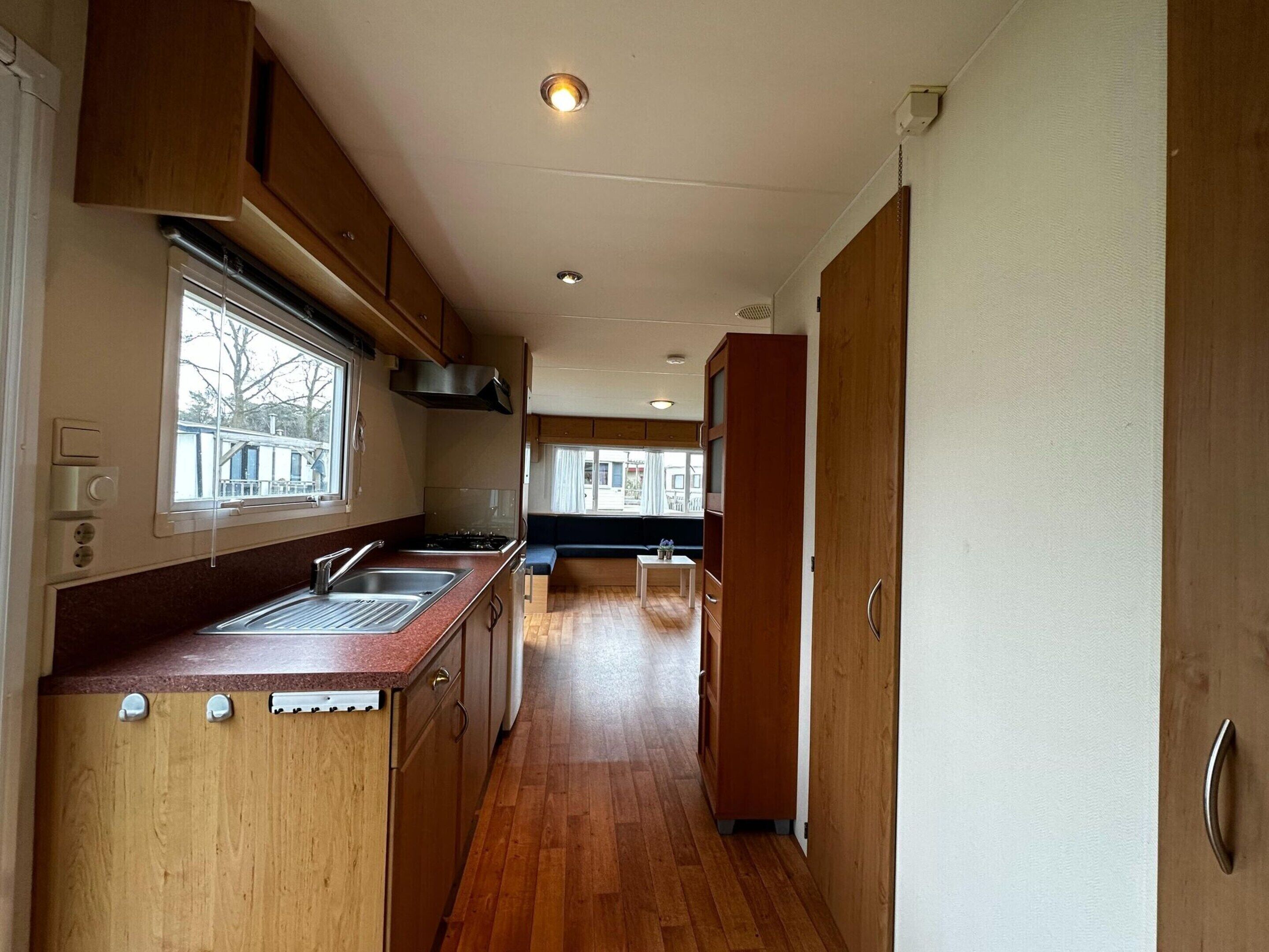 A narrow kitchen with wooden cabinets, a stainless steel sink, and a window overlooking a building.