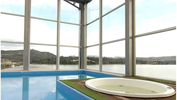 A large indoor swimming pool with a hot tub, surrounded by large windows offering a view of a lake and hills. - Apartamento Familiar de Lujo al Lado del Lago (Paipa)