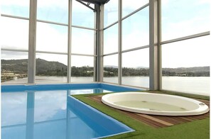 Une grande piscine intérieure avec un jacuzzi, entourée de larges baies vitrées donnant sur un lac et des collines.
