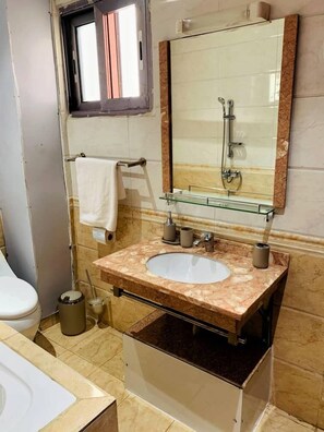 Un baño con lavamanos de mármol, estante de vidrio, toallero y espejo con ducha.