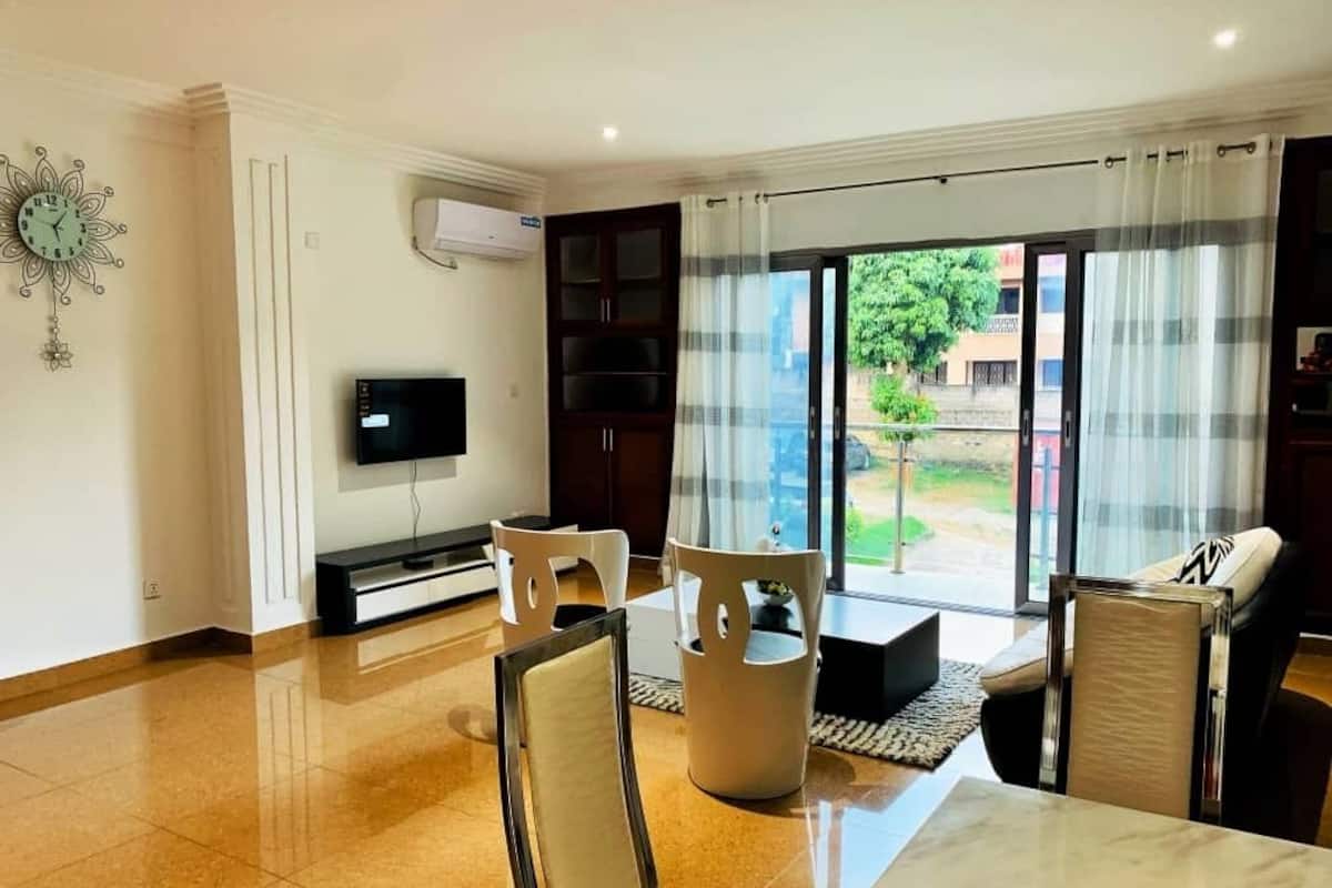 A modern living room with a flat-screen TV, a dining table, and chairs. There is a sliding glass door leading to a balcony with a view of the outside.
