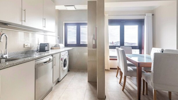 A modern kitchen with stainless steel appliances, a dishwasher, and a washing machine. There is a dining area with a table and chairs, and a large window providing a view of the city.