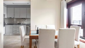 A dining area with a table and chairs, a kitchen in the background, and a window with curtains.