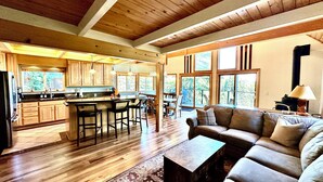 A spacious living area with a large sofa and a coffee table, a kitchen with wooden cabinets and a bar, and large windows offering views of the outdoors. - Tahoe Treetop Escape 4BR Near Trails & Donner Lake (Truckee,)
