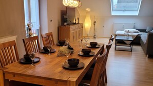 A dining area with a long wooden table set for six, flanked by wooden chairs. A cabinet with decorative items and a floor lamp are visible in the background.